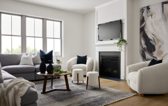 Modern living room with a gray sectional sofa, round wooden coffee table, two white armchairs, abstract wall art, and a fireplace with a TV above it. Large windows let in natural light, and plants add greenery.