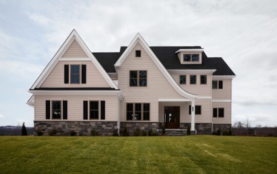 A large, modern two-story house with light beige siding, dark-framed windows, and a stone foundation sits on a well-manicured lawn under a cloudy sky.