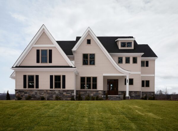A large, modern two-story house with light beige siding, dark-framed windows, and a stone foundation sits on a well-manicured lawn under a cloudy sky.