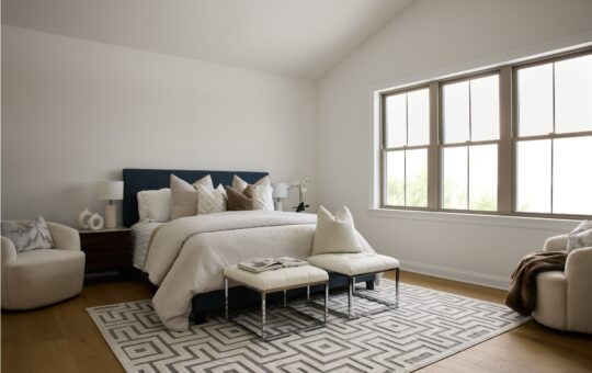 A modern, minimalistic bedroom with a neatly made bed, neutral bedding, multiple pillows, two white armchairs, large windows, a geometric area rug, two benches at the foot of the bed, and wooden floors.