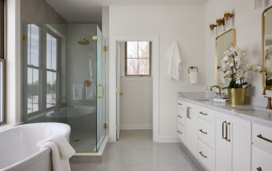 A modern bathroom with a glass shower, freestanding bathtub, double sinks with gold fixtures, white cabinetry, and a large mirror. A towel hangs on the tub and fresh white orchids are on the countertop.