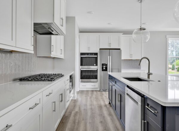 Modern kitchen with white cabinets, stainless steel appliances, a gas cooktop, built-in oven, gray refrigerator, large island with sink and dishwasher, pendant lights, and LVP flooring. Bright natural light from window.