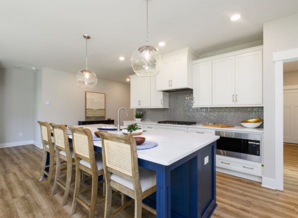 Modern kitchen with a blue island, white cabinets, wood flooring, rattan bar stools, pendant lights, and stainless steel appliances; a decorative tray and plant sit on the island countertop.