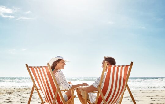 A couple sits in orange-and-white striped deck chairs on a sandy beach, holding hands and smiling at each other under a bright sunny sky with the ocean in the background.