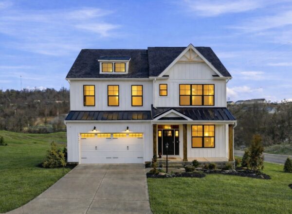 A two-story modern farmhouse with white siding, large windows glowing warmly, a black metal roof, double garage doors, and a covered porch, set on a spacious green lawn with a scenic background at dusk.