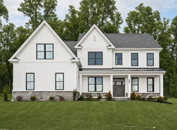 A modern two-story white house with dark framed windows, a gray shingled roof, stone accents, and a manicured lawn, set in front of tall green trees under a blue sky.