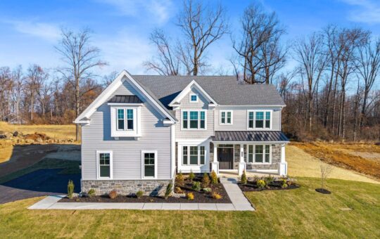Two-story modern house with gray siding, white trim, and black roof, set on a large grassy lawn with a paved walkway and driveway. Sparse landscaping and leafless trees in the background under a blue sky.