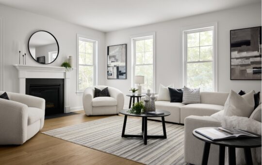 A modern living room with white walls and furniture, black accents, a round mirror above a fireplace, large windows letting in natural light, abstract artwork, and a striped rug on a wooden floor.