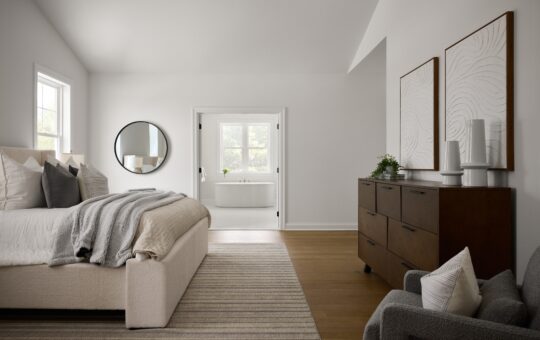 A modern, minimalist bedroom with neutral tones, a large upholstered bed, a striped rug, a wooden dresser with decor, and a round mirror. An ensuite bathroom with a tub is visible through double doors.