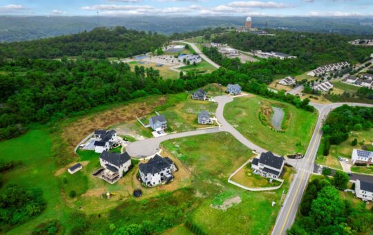 Aerial view of a suburban neighborhood with several large houses under construction, surrounded by green hills, trees, and distant buildings under a partly cloudy sky.
