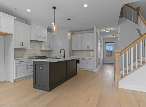 Modern kitchen with white cabinets, a black island with sink, pendant lights, light wood floors, stainless steel appliances, subway tile backsplash, and a staircase with wood railings—showcasing Foxlane Homes Chalfont craftsmanship.