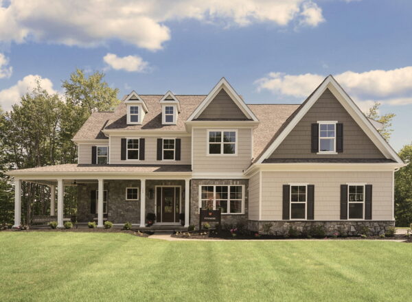 Two-story suburban house with gray siding, white trim, multiple gables, and black shutters. A porch with a swing is on the left. Lush green lawn and trees are in the background under a blue sky with clouds.
