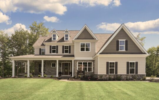 A large two-story suburban house with gray siding, white trim, and black shutters sits on a manicured lawn under a blue sky with clouds. Trees line the background.
