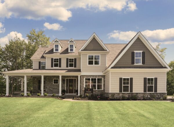 A large two-story suburban house with gray siding, white trim, and black shutters sits on a manicured lawn under a blue sky with clouds. Trees line the background.