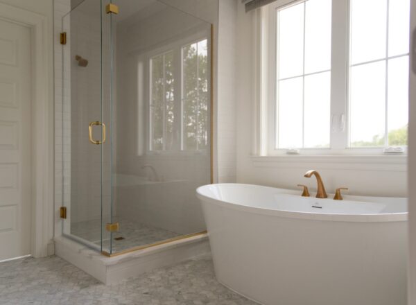 A modern bathroom with a glass shower, gold fixtures, a white freestanding bathtub, and large windows letting in natural light.