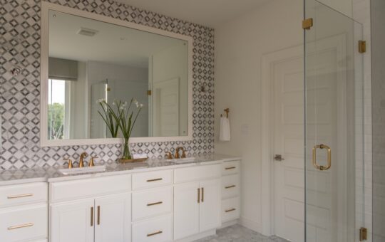 Modern bathroom with white cabinets, dual sinks, gold fixtures, a large mirror, geometric patterned wall tiles, a glass shower door, and a vase with white flowers on the counter.