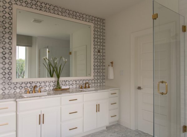 Modern bathroom with white cabinets, dual sinks, gold fixtures, a large mirror, geometric patterned wall tiles, a glass shower door, and a vase with white flowers on the counter.