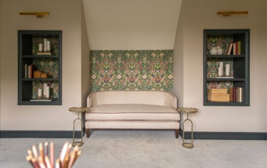 A small cream sofa sits between two built-in shelves with books and decor, against a wall with floral green wallpaper. Two round gold side tables flank the sofa, and soft light fills the carpeted room.
