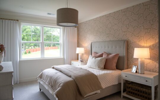 A cozy bedroom with a large window, upholstered bed with neutral bedding, two bedside tables with lamps, and patterned wallpaper. Natural light brightens the room.