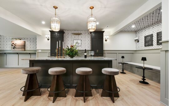 Modern kitchen with a large island, four round stools, dark cabinets, two pendant lights, patterned backsplash, and a long bench seating area with small black lamps along the right wall.