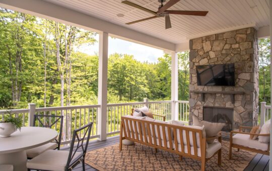 Covered porch with outdoor seating, stone fireplace, wall-mounted TV, ceiling fan, a dining table, and views of green trees and lawn beyond the white railing.
