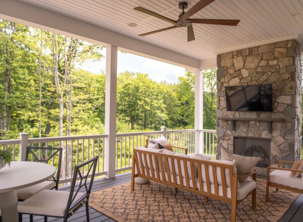 Covered porch with outdoor seating, stone fireplace, wall-mounted TV, ceiling fan, a dining table, and views of green trees and lawn beyond the white railing.