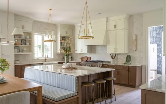 Modern kitchen with white cabinets, wood accents, a large island with striped bench seating, three pendant lights, and gold fixtures. Sunlight streams in, highlighting a clean, bright, and airy atmosphere.