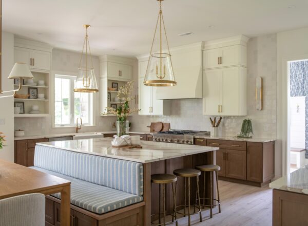 Modern kitchen with white cabinets, wood accents, a large island with striped bench seating, three pendant lights, and gold fixtures. Sunlight streams in, highlighting a clean, bright, and airy atmosphere.