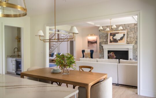 Open-concept dining and living area with wooden table, beige chairs, chandelier, neutral sofas, stone fireplace, modern art, and plants. Light tones and natural elements create a cozy, elegant space.