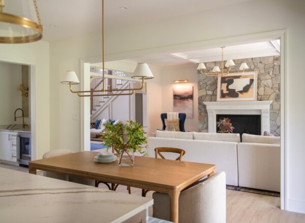 Open-concept dining and living area with wooden table, beige chairs, chandelier, neutral sofas, stone fireplace, modern art, and plants. Light tones and natural elements create a cozy, elegant space.