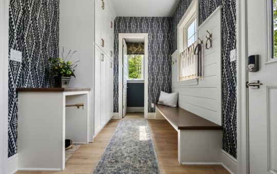 A bright mudroom with patterned wallpaper, white built-ins, a wooden bench with hooks, a window, flowers on a shelf, and a gray runner rug on light wood flooring. An open door reveals greenery outside.