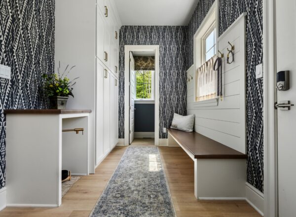 A bright mudroom with patterned wallpaper, white built-ins, a wooden bench with hooks, a window, flowers on a shelf, and a gray runner rug on light wood flooring. An open door reveals greenery outside.