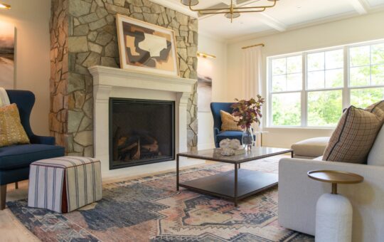 Modern living room with stone fireplace, abstract art, blue armchairs, cushioned ottoman, patterned rug, coffee table with decor, and large windows letting in natural light.