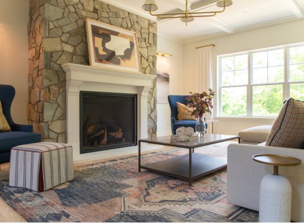 Modern living room with stone fireplace, abstract art, blue armchairs, cushioned ottoman, patterned rug, coffee table with decor, and large windows letting in natural light.