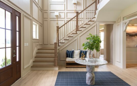 Elegant entryway with a staircase, wood flooring, a round marble table with a plant, a patterned rug, and a large windowed door letting in natural light.