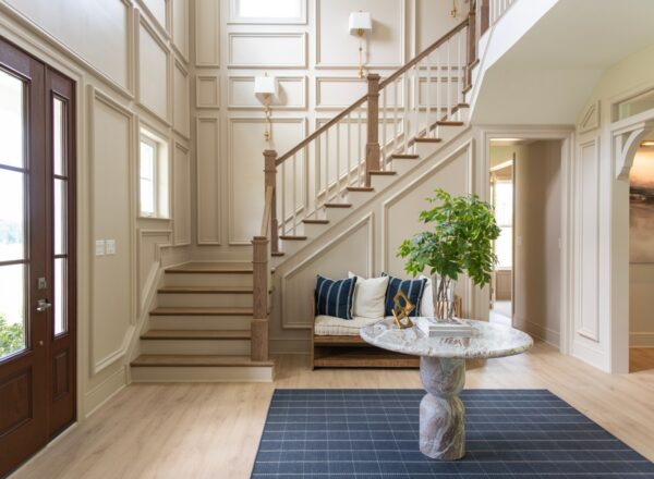 Elegant entryway with a staircase, wood flooring, a round marble table with a plant, a patterned rug, and a large windowed door letting in natural light.