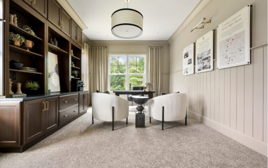 Modern home office with built-in dark wood shelves, two white chairs, a desk, large window with beige curtains, and wall art. Neutral tones and carpeted floor create a bright, elegant space.
