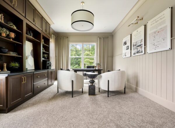 Modern home office with built-in dark wood shelves, two white chairs, a desk, large window with beige curtains, and wall art. Neutral tones and carpeted floor create a bright, elegant space.