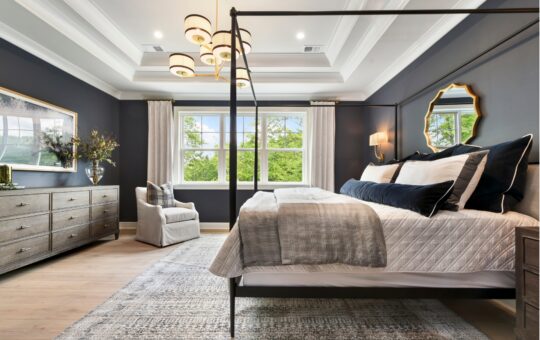 Elegant bedroom with a canopy bed, gray walls, large windows, a dresser, armchair, modern chandelier, gold-framed mirrors, and light-colored rug. Natural light fills the room, creating a cozy and inviting atmosphere.