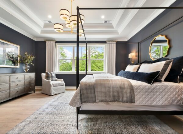 Elegant bedroom with a canopy bed, gray walls, large windows, a dresser, armchair, modern chandelier, gold-framed mirrors, and light-colored rug. Natural light fills the room, creating a cozy and inviting atmosphere.