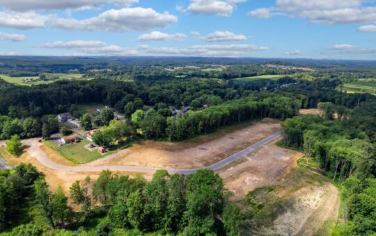 Aerial view of a partially cleared land area surrounded by dense trees, with a winding road cutting through. Nearby are houses, open fields, and a mix of forested areas under a blue sky with scattered clouds.