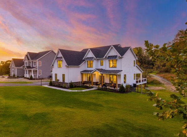 A large white modern house with glowing windows sits on a spacious green lawn at sunset, with another similar house nearby under a colorful pink and blue sky.