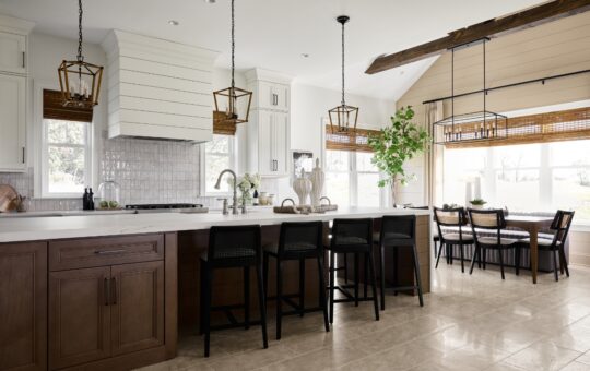 Modern kitchen with wood cabinets, a large island with black barstools, pendant lights, a shiplap vent hood, and a dining nook with a table and chairs by large windows. A potted plant sits near the dining area.