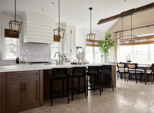 Modern kitchen with wood cabinets, a large island with black barstools, pendant lights, a shiplap vent hood, and a dining nook with a table and chairs by large windows. A potted plant sits near the dining area.