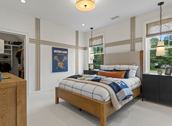 A modern bedroom with a wooden bed, plaid bedding, and sports-themed decor. Two pendant lights hang by windows with woven shades. A dresser and open closet are also visible. The room is bright with neutral walls and carpet.