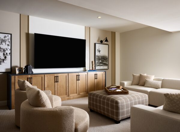 Modern living room with beige walls and carpet, featuring a large flat-screen TV on a wooden cabinet, abstract artwork, cream sofas, round chairs, a plaid ottoman, and decorative pillows.