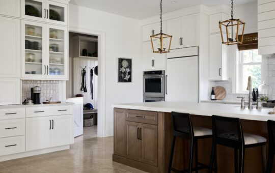 Modern kitchen with white cabinets, a large island with wooden base, black chairs, pendant lights, built-in oven, and a walk-in pantry. Neutral tones, ample natural light, and minimalistic decor.