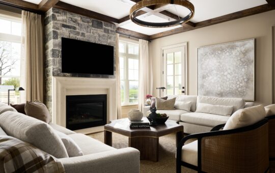 A cozy living room with cream sofas, a stone fireplace, a large TV, a circular chandelier, and a modern coffee table. Light flows in through tall windows and a glass door, highlighting neutral tones and a large abstract painting.