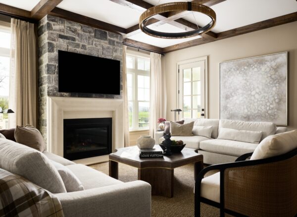 A cozy living room with cream sofas, a stone fireplace, a large TV, a circular chandelier, and a modern coffee table. Light flows in through tall windows and a glass door, highlighting neutral tones and a large abstract painting.