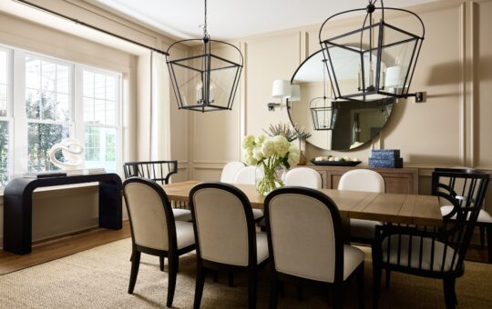 Elegant dining room with a wooden table, eight cushioned chairs, two large black lantern pendant lights, a round mirror, sideboard, white flowers in a vase, and natural light from large windows.